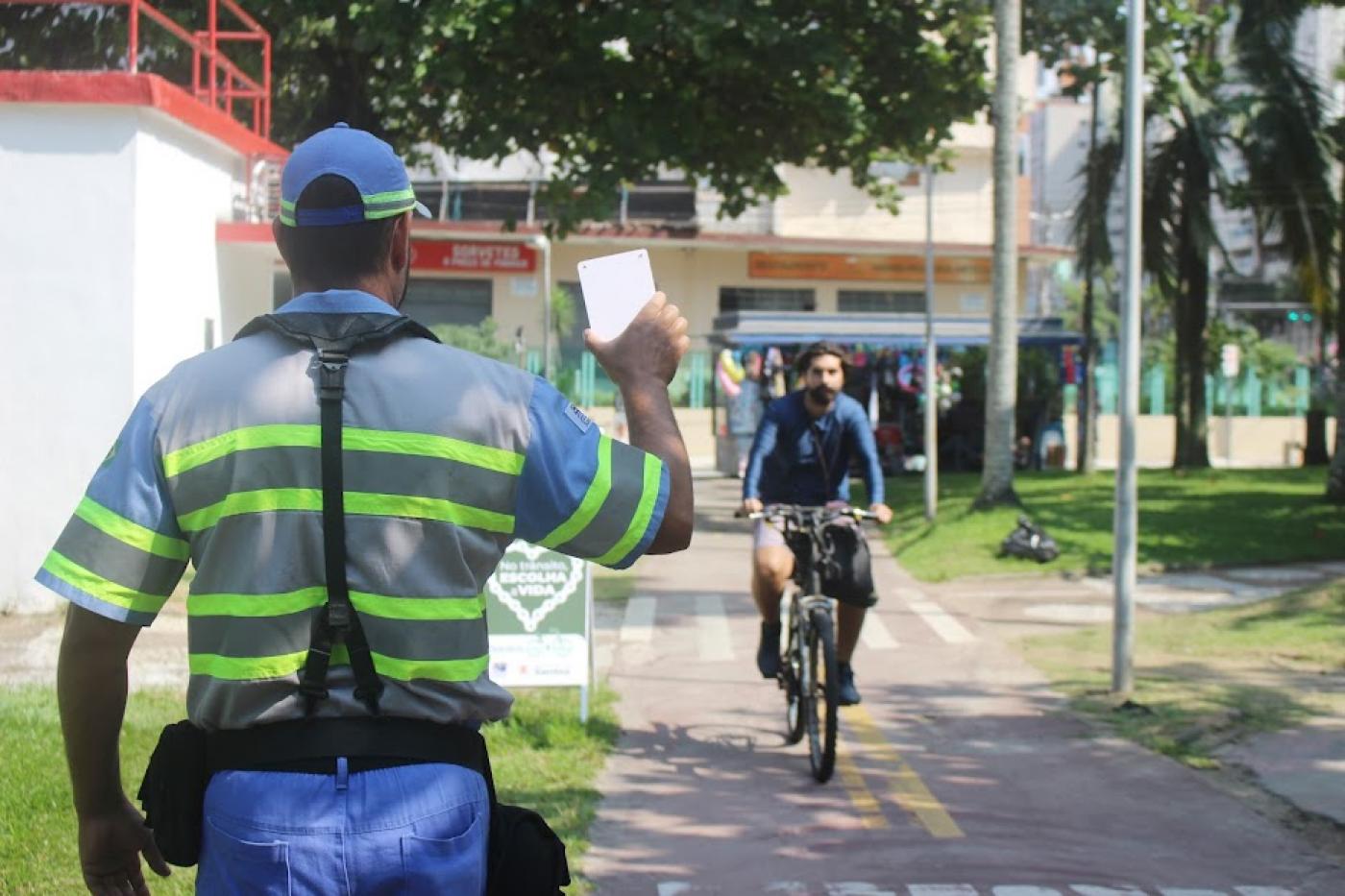 agente de trânsito está de costas segurando uma plaquinha. Ele está ao lado da ciclovia, por onde passam ciclistas. #paratodosverem 