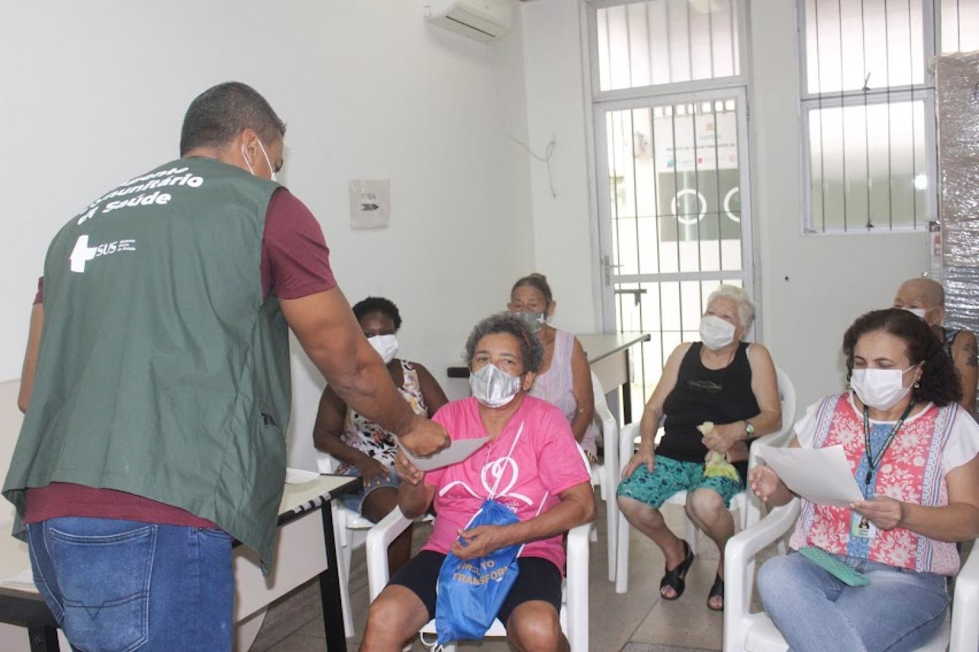 homem está entregando folheto para mulheres sentadas em sala de policlínica. #paratodosverem