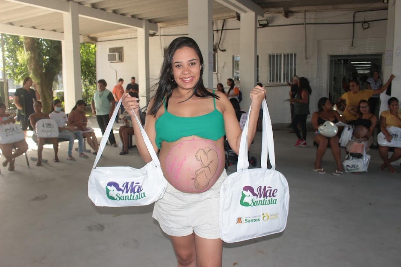 gestante com a barriga de fora segura duas sacolas do mãe santista. #paratodosverem