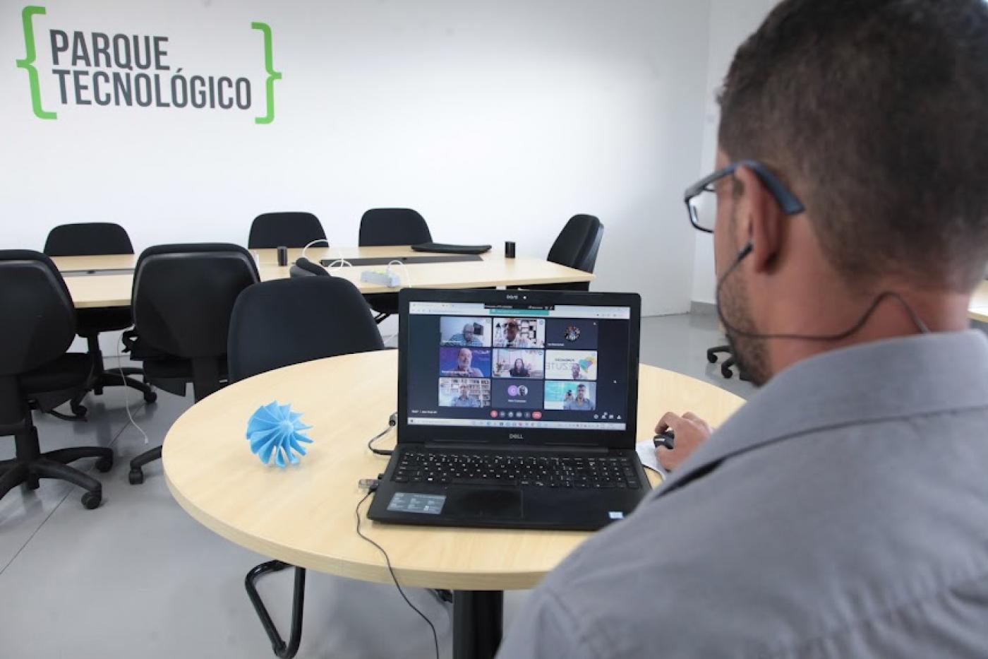 homem está de costas para a foto e diante de um monitor de computador que está sobre uma mesa. Ao lado do monitor, um pequeno protótipo de gerador de energia.  #paratodosverem