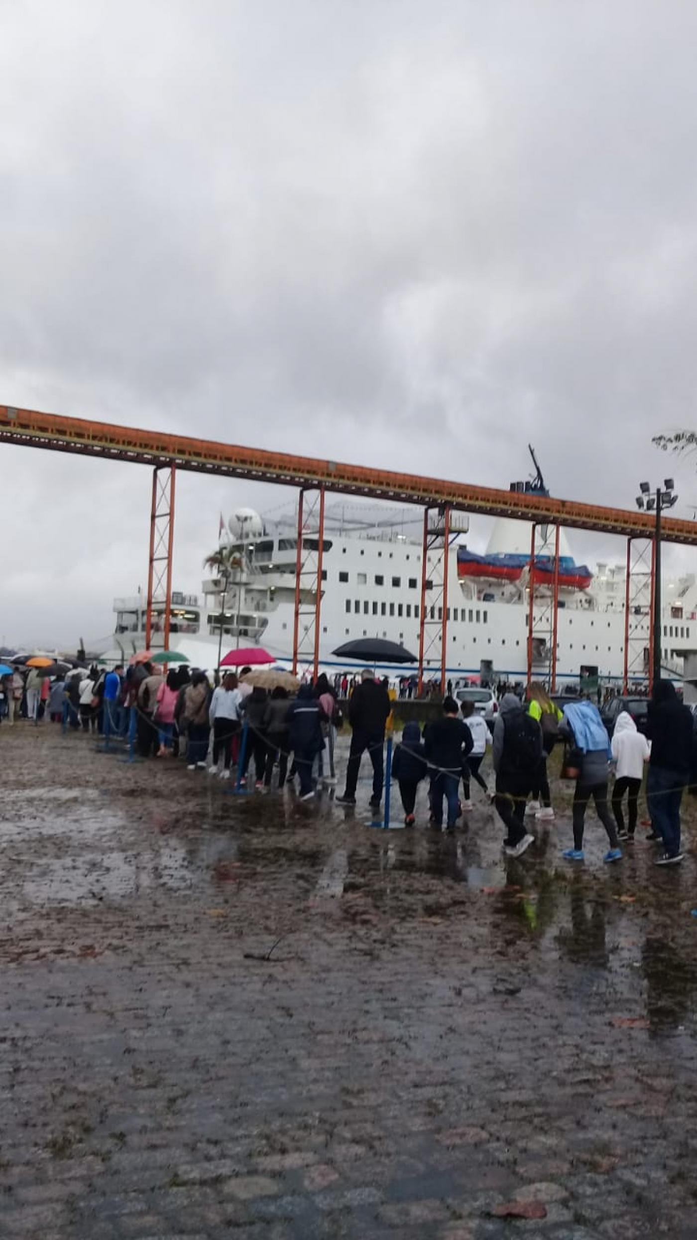 Fila de pessoas no cais para entrar no navio. Dia está chuvoso. 