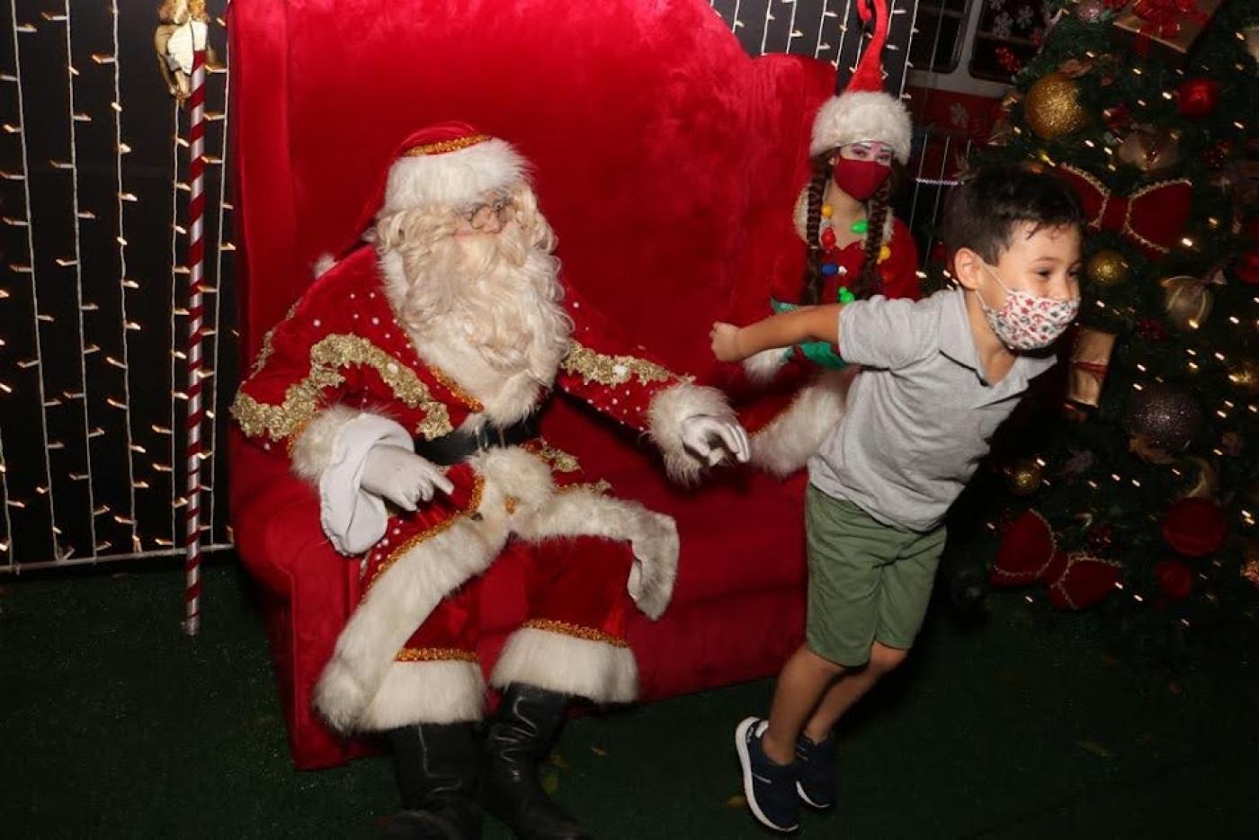 menino sai da cadeira onde está papai noel pulando de alegria. A mamãe noel e uma árvore de natal estão ao fundo. #paratodosverem