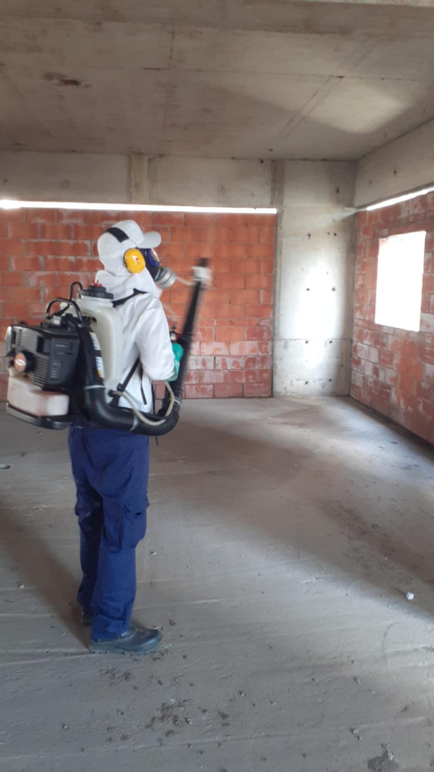 homem coberto por uniforme, que inclui capacete, máscara especial, capa, calça e botas, faz aplicação de inseticida a partir de equipamento tipo mochila nas costas. #paratodosverem