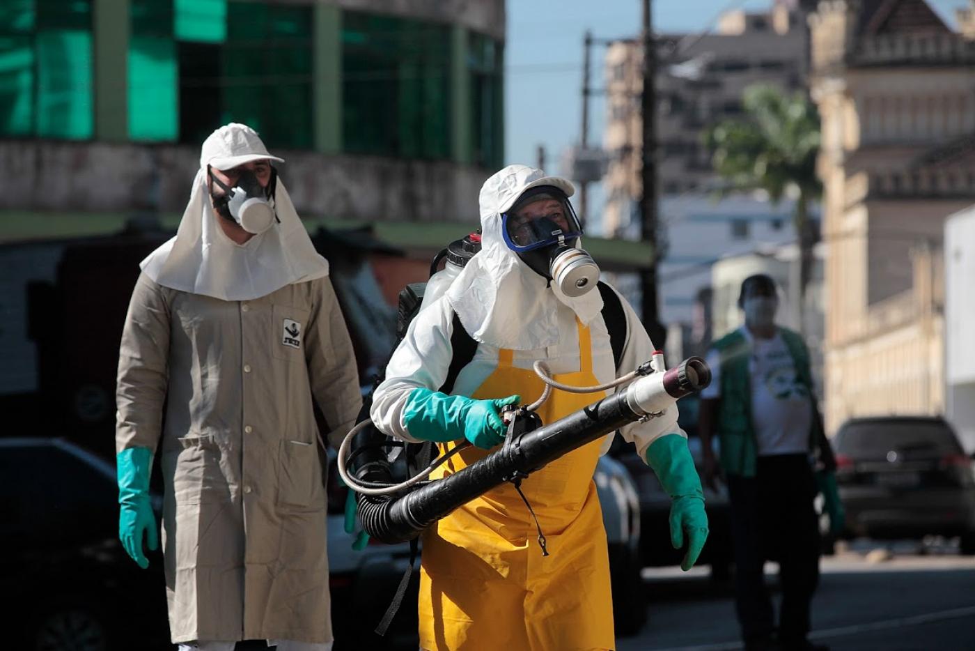 Dois homens paramentados com máscaras e luvas. Um deles carrega um equipamento para aplicar inseticida. #Paratodosverem