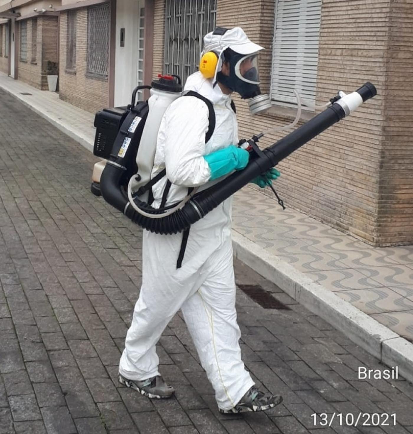 homem está usando equipamento de nebulização do tipo costal, com uma espécie de mochila nas costas ligada a um tubo similar ao de um aspirador de pó. Ele está vestido com macacão, mascara própria para respirar e usa proteção no ouvido. #paratodosverem