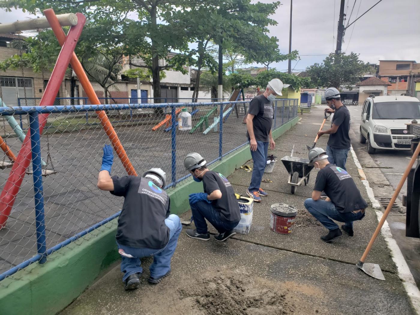 homens trabalham em obra em calçada ao lado de play ground com alambrado. São cinco pessoas, três estão agachadas junto a latas de tinta, pá e carrinho de mãos. Eles usam capacete. #paratodosverem