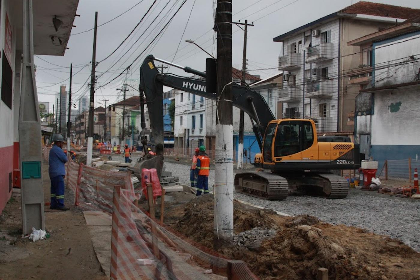 rua com obras. Retroescavadeira em movimento e homens trabalhando. há terra nos canteiros e a pista está coberta  por pedras. #paratodosverem