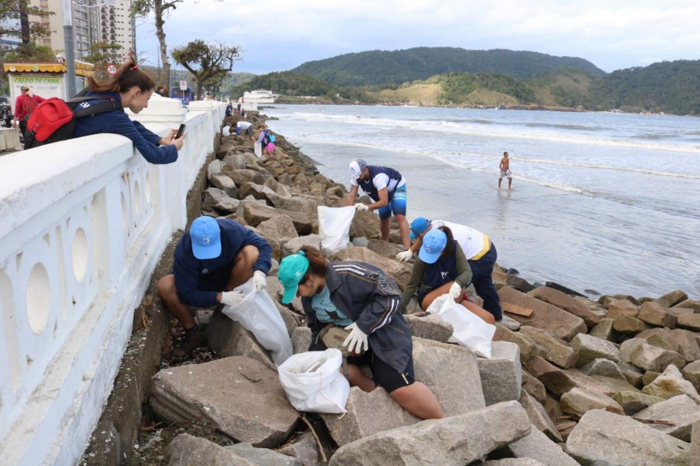 voluntários estão junto às pedras da mureta da ponta da praia, como mar ao fundo. Eles recolhem microlixo. #paratodosverem