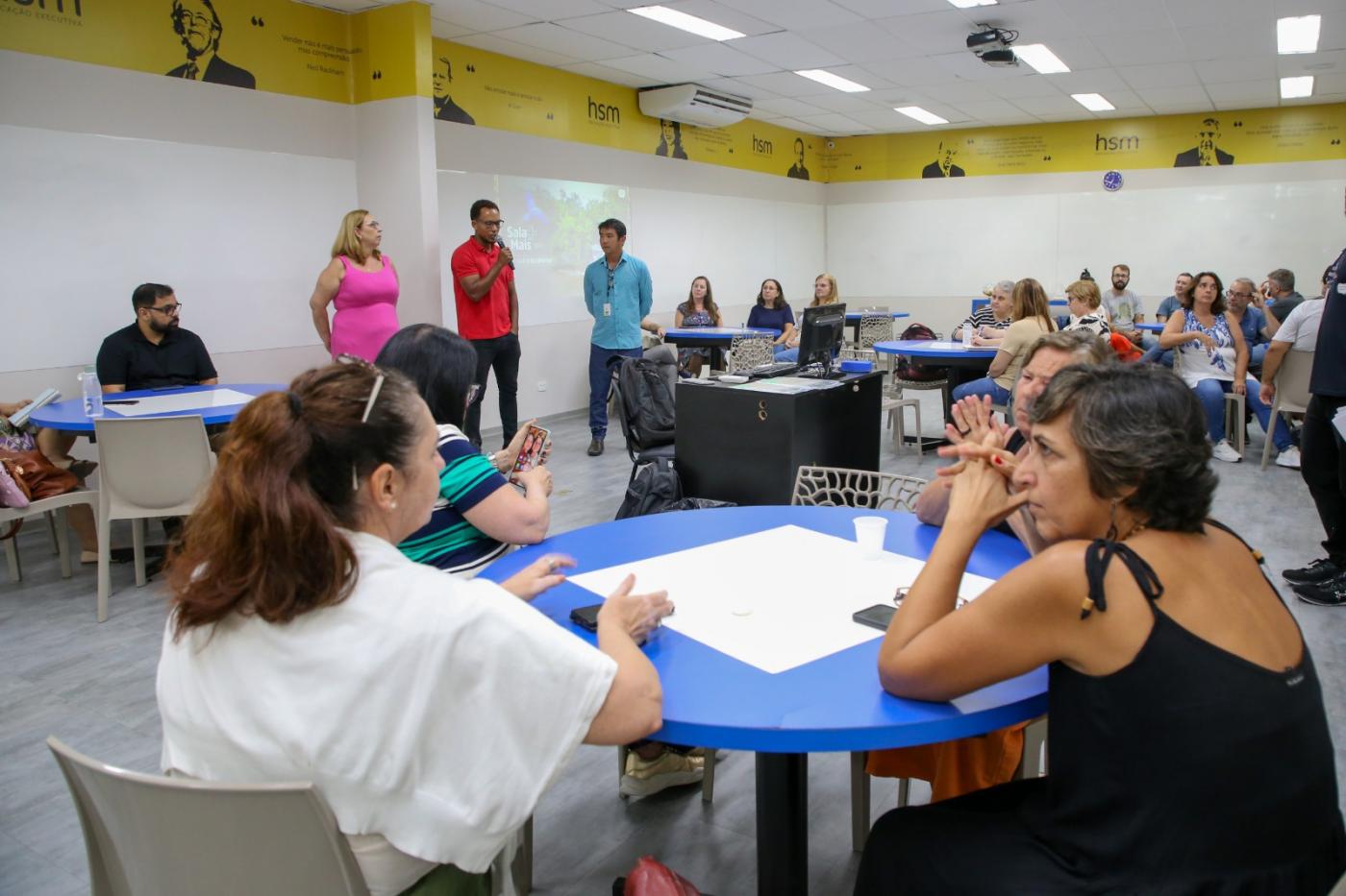 em primeiro plano, mesa com professoras sentadas. Ao fundo, três pessoas em pé, sendo uma delas falando ao microfone. Há outros professores sentados em torno. #paratodosverem 