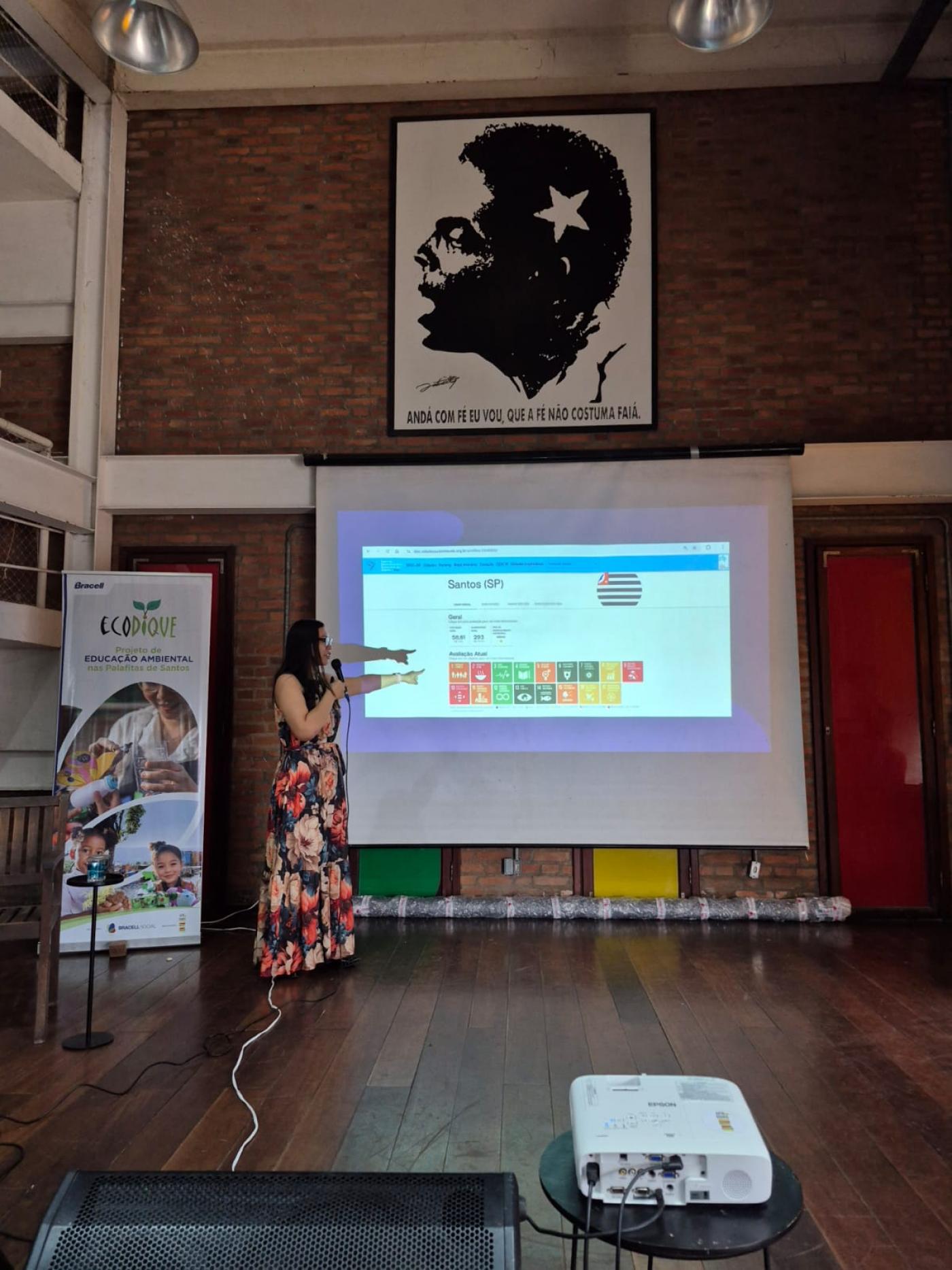 mulher dá palestra apontando em telão #paratodosverem