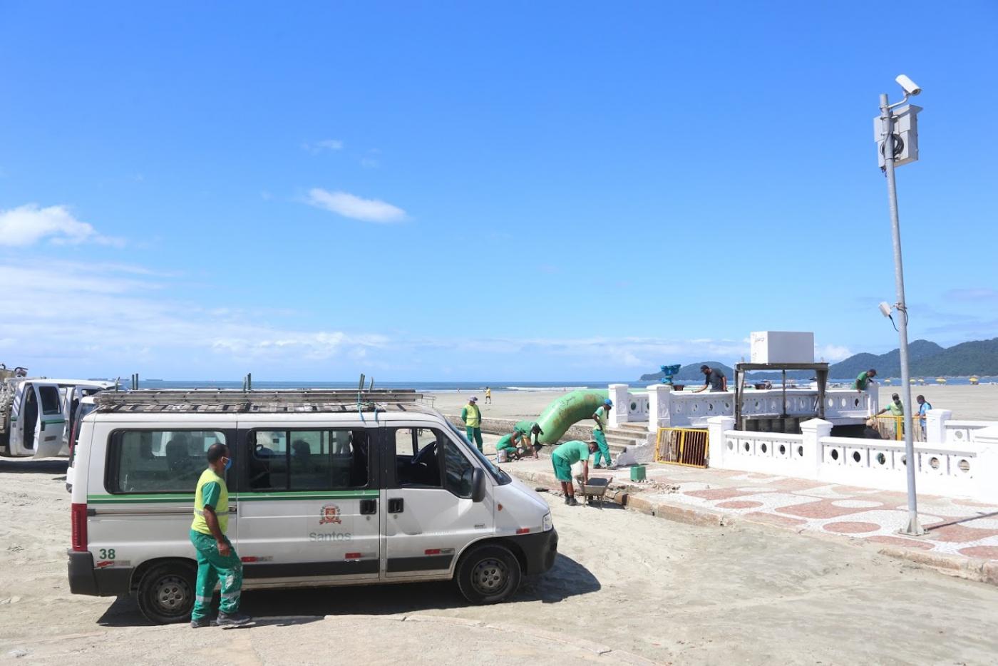 Homens trabalham próximo ao canal, fazendo manutenção. Há um carrinho de mão e um veículo de apoio na areia.#Paratodosverem