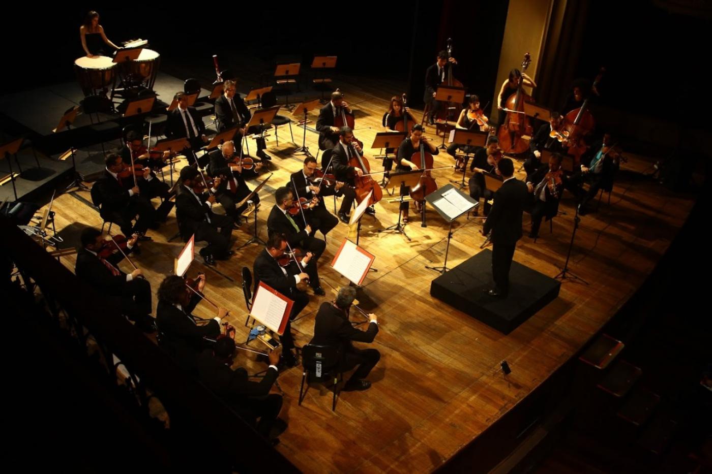 Músicos da orquestra tocam em palco com maestro à frente. #paratodosverem