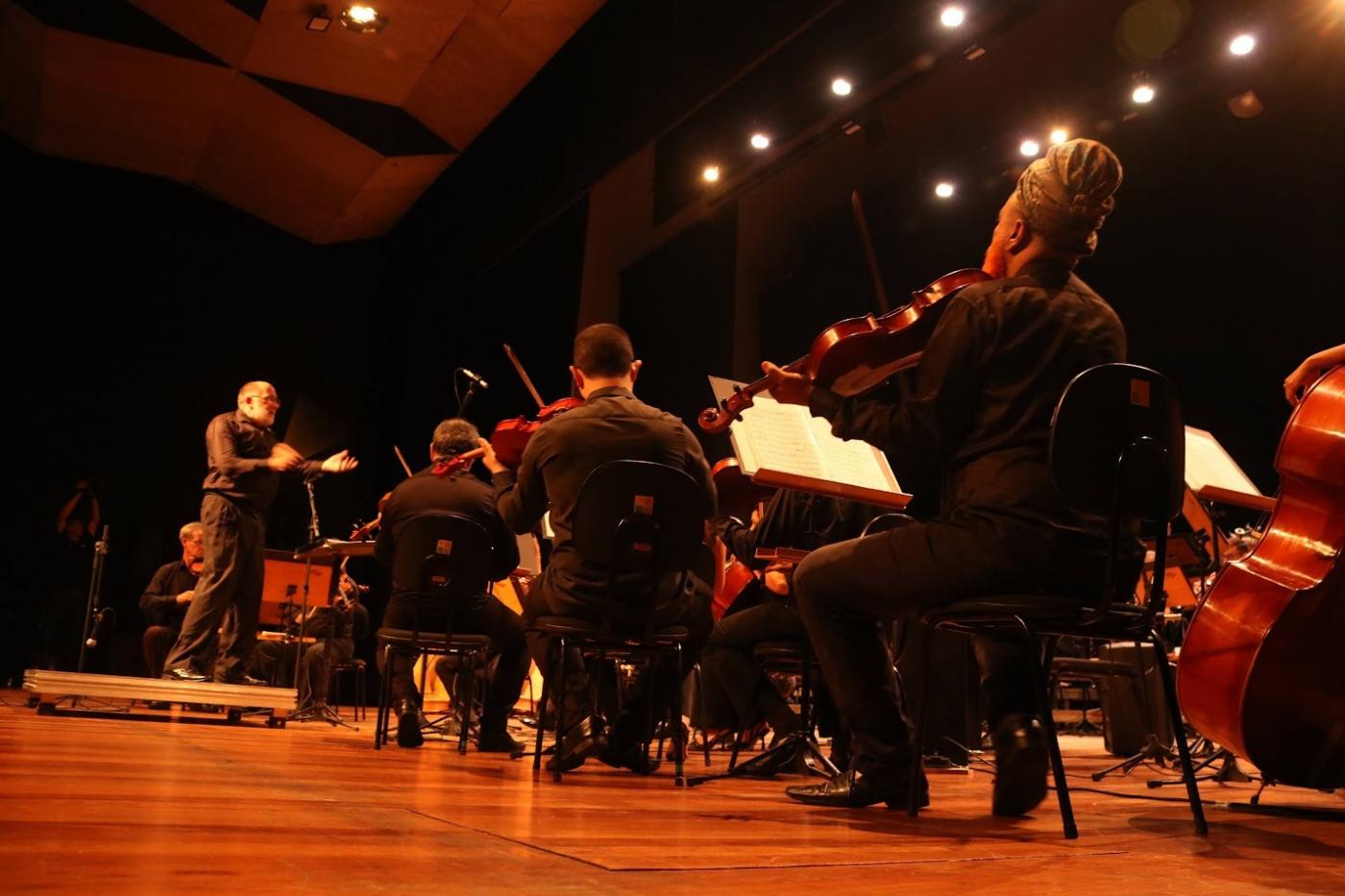 músicos sentados tocando com maestro ao fundo num teatro. #paratodosverem 