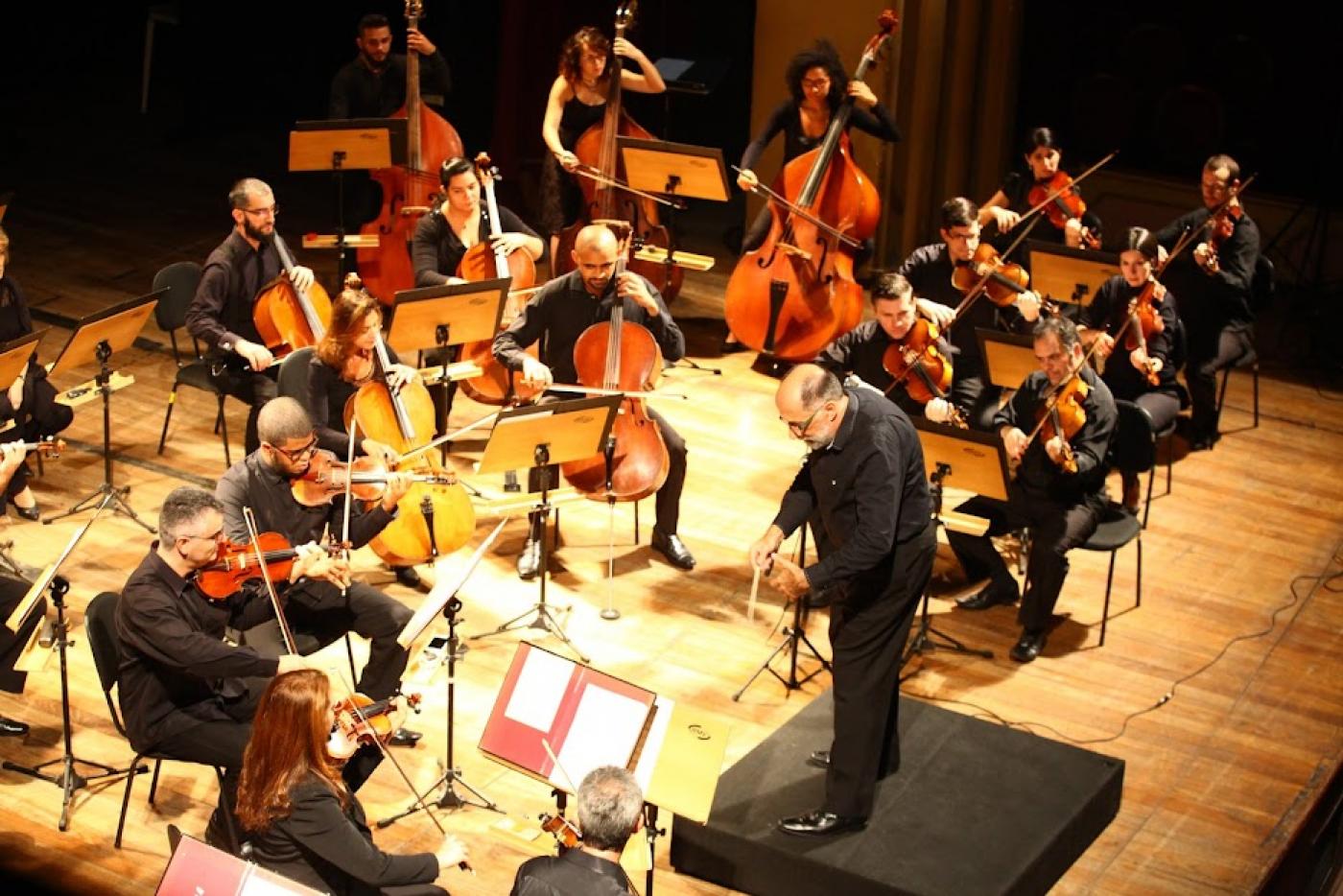 maestro está à frente da orquestra. Todos estão no palco. #paratodosverem