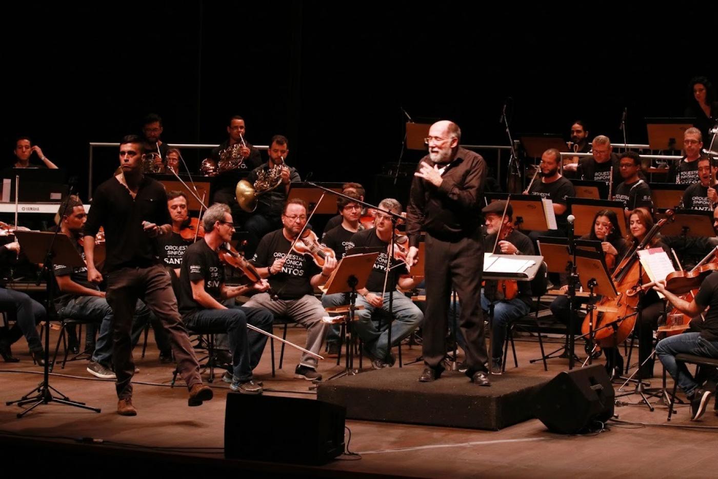 maestro está à frente da orquestra. Ele olha para a frente. Músicos estão atrás. #paratodosverem