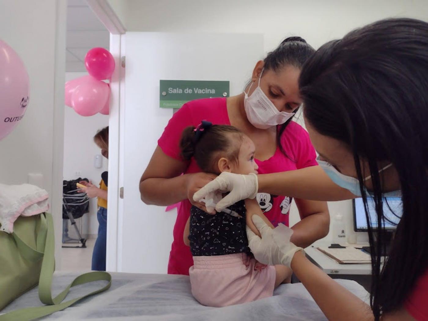 Criança recebe vacina com mãe segurando #paratodosverem