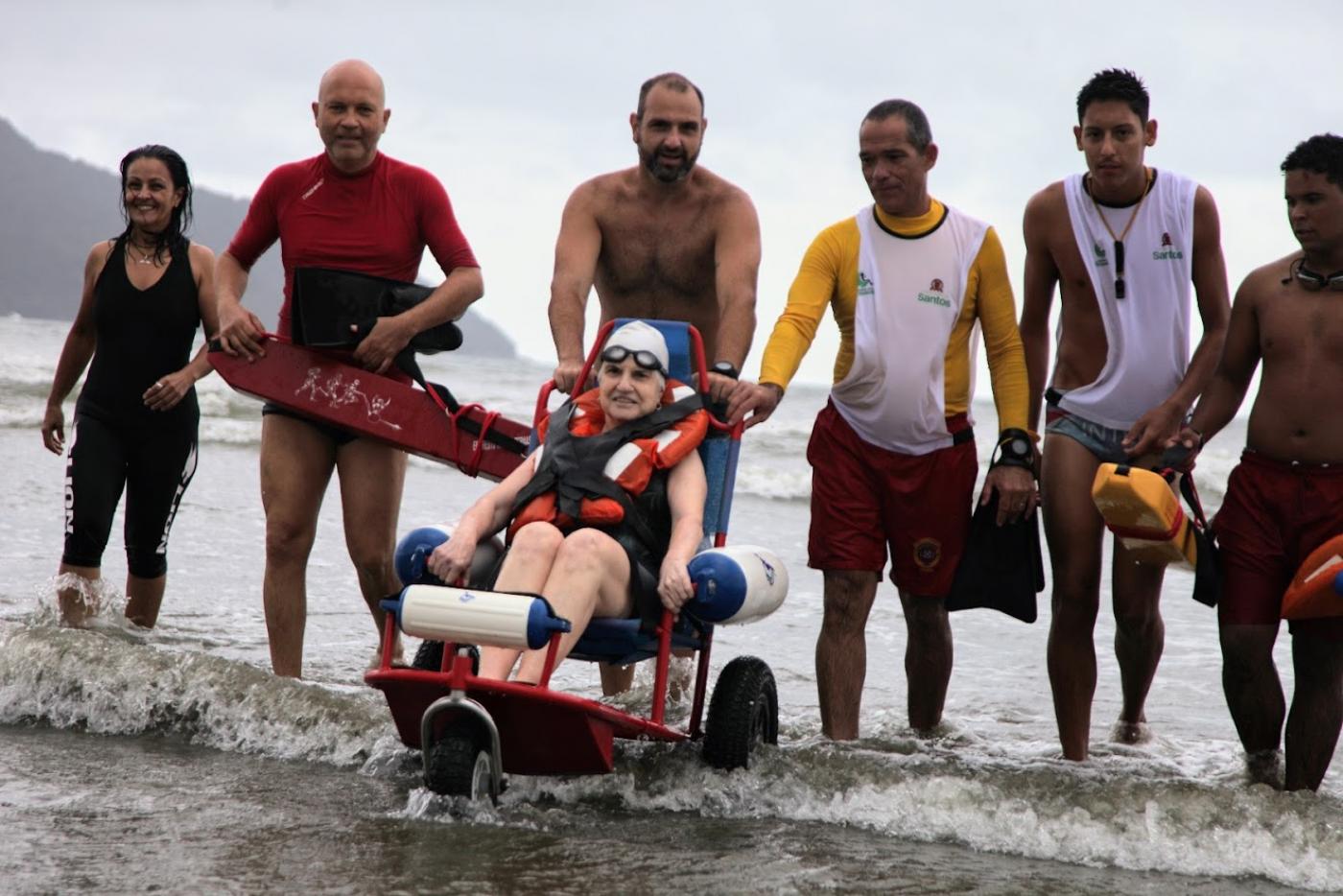 Mulher está sentada em cadeira anfíbia, na beira d'água. Ela está ladeada por um grupo de seis pessoas