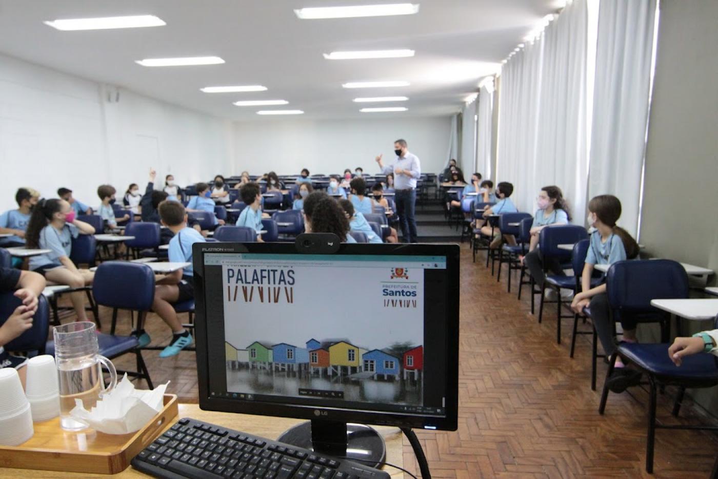 Computador com tela sobre projeto e crianças e prefeito ao fundo #paratodosverem