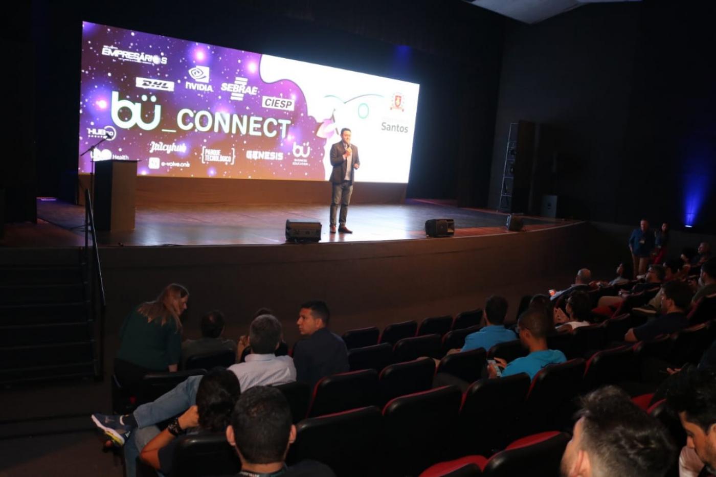 homem está falando no palco diante um telão. Pessoas estão sentadas na plateia. #paratodosverem