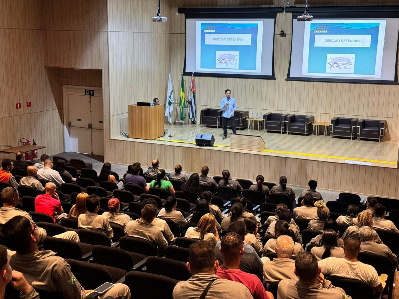 auditório lotado de pessoas assistindo palestra de uma pessoa no palco. Há telões com mensagens ao fundo. #paratodosverem 
