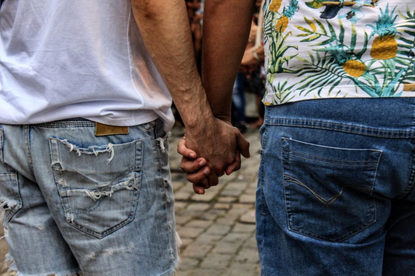 Dois homens de mãos dadas. Eles estão de costas. Imagem em close sobre as mãos. Eles caminham pela rua. #Pracegover
