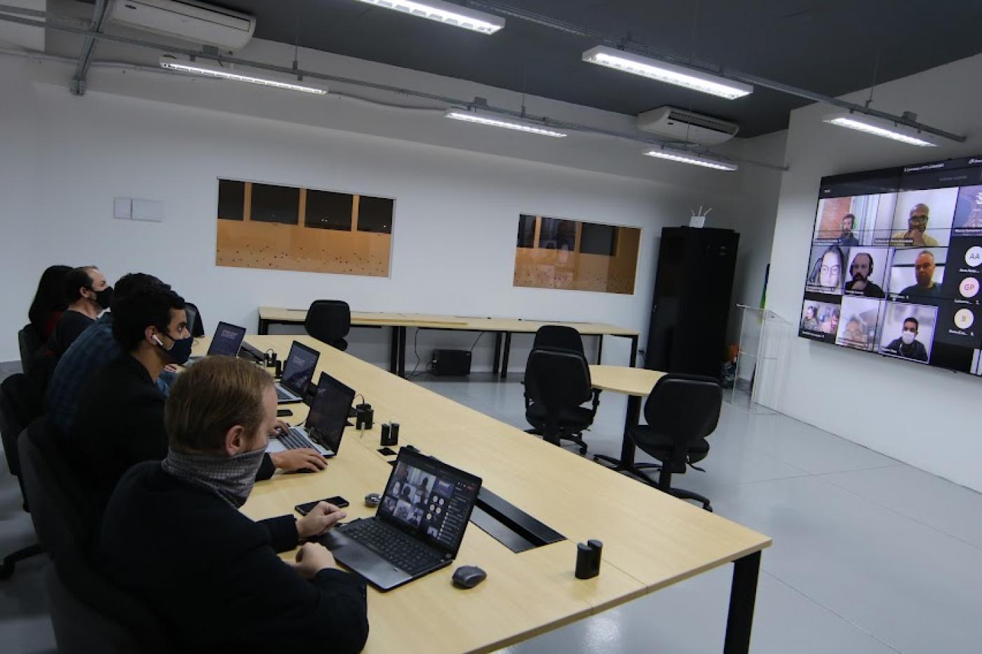 Pessoas estão sentadas diante de seus laptops aberto e assistindo a uma apresentação em telão. #paratodosverem