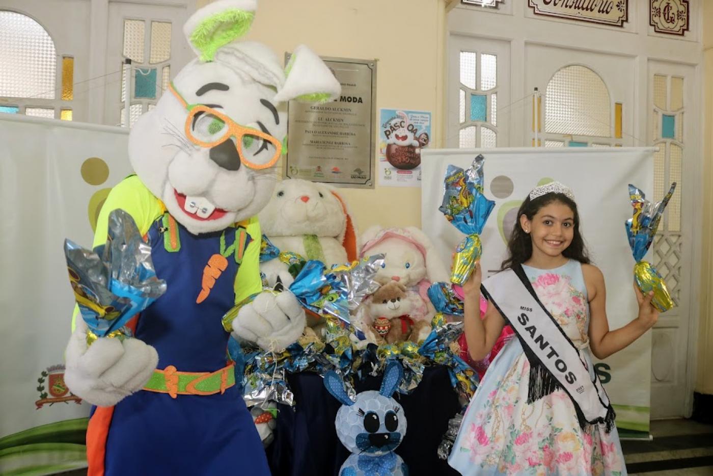 Pessoa vestida com fantasia de coelho e a miss santos mirim estão à frente de mesas com bonecos e ovos de chocolate. Eles dois também seguram ovos nas quatro mãos. #paratodosverem