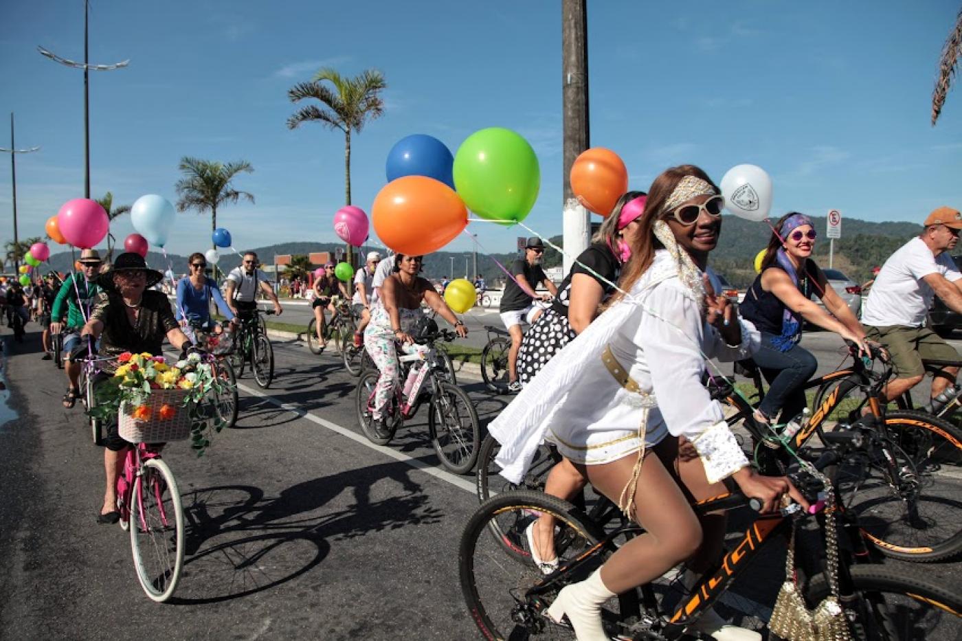 pessoas vestidas com roupas antigas e segurando balões de gás estão montadas em suas bikes e pedalando na avenida da orla. #paratodosverem