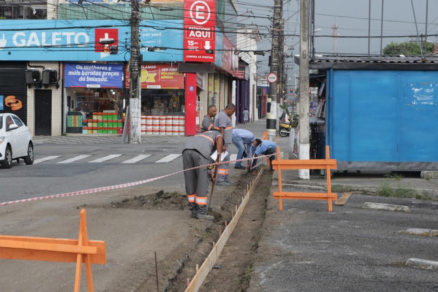 Trecho está delimitado por cavalete e fita. Operários está manuseando enxadas junto à guia. #Paratodosverem