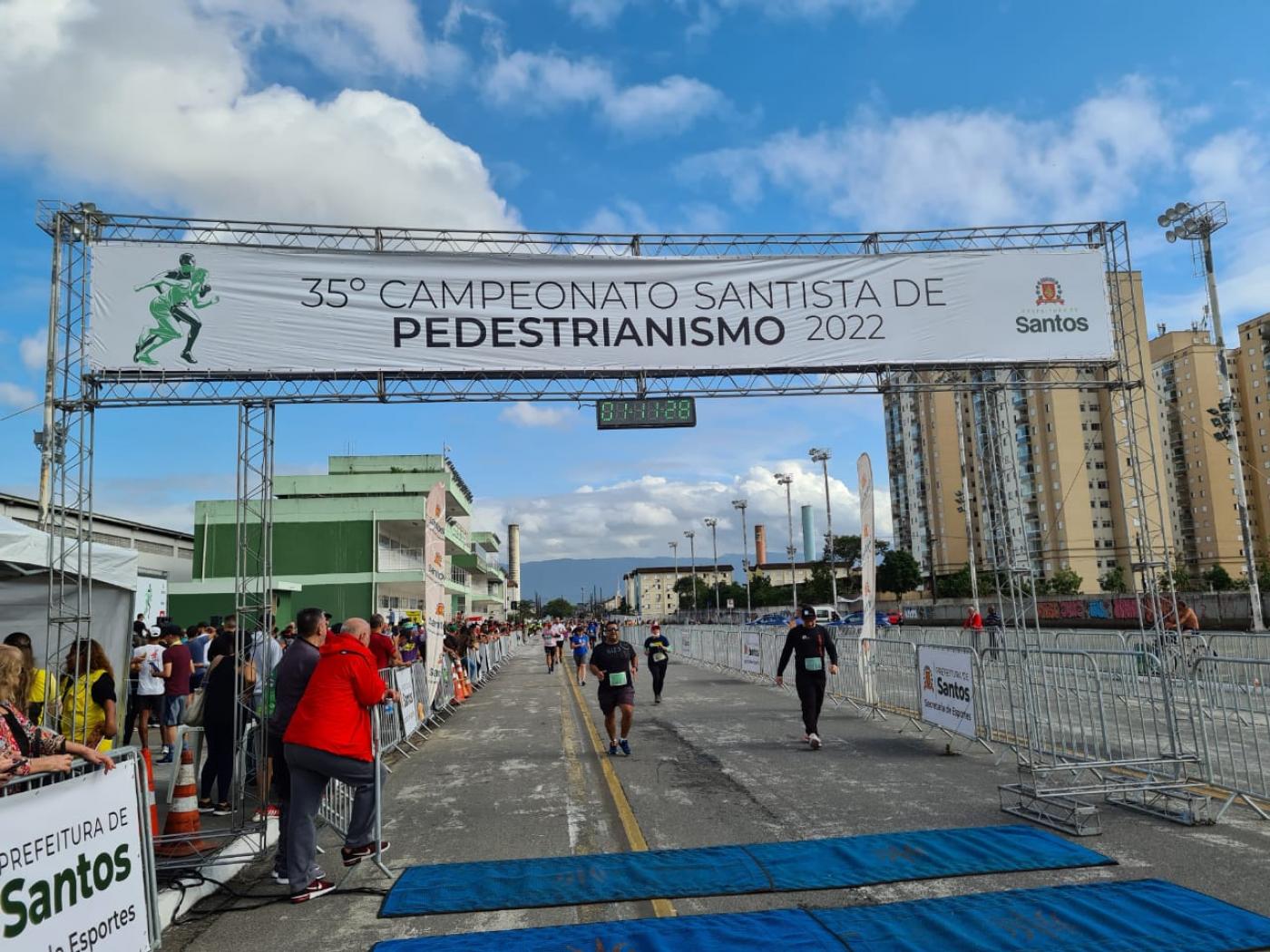 atletas estão chegando ao final da prova. Há um pórtico no alto onde se lê 35º Campeonato Santista de Pedestrianismo. #paratodosverem