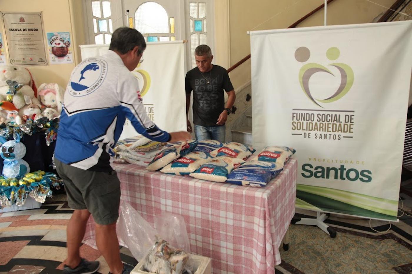 mesa com pacotes de alimentos e um homem colocando mais pacotes. Ao fundo, um banner do fundo social. #paratodosverem