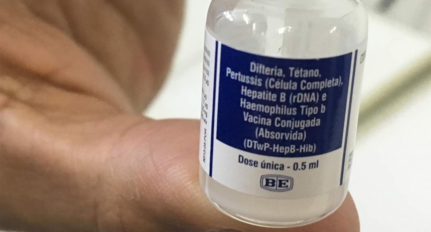 Imagem em close mostra mão segurando frasco com vacina pentavalente. #Pracegover