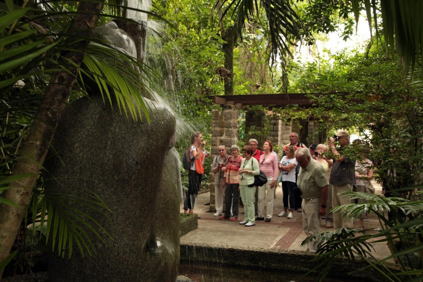 Grupo de turistas está na entrada do orquidário. Em primeiro plano está a ninfa Náiade, de costas. #Paratodosverem