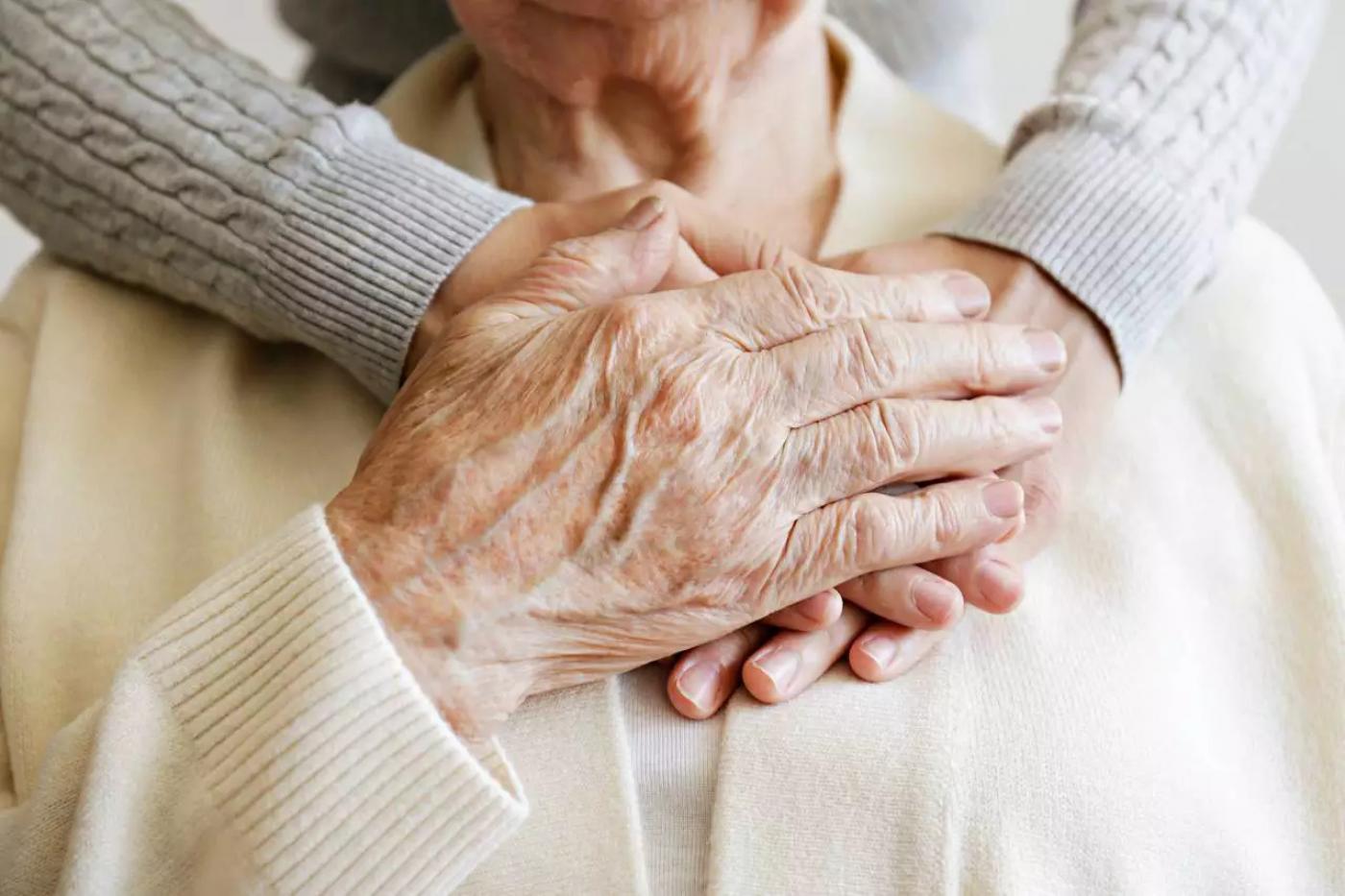 mão de pessoa idosa está sobre duas mãos de pessoa mais jovem que a abraçam por trás, na altura do peito. A imagem está com close nas mãos. #paratodosverem 