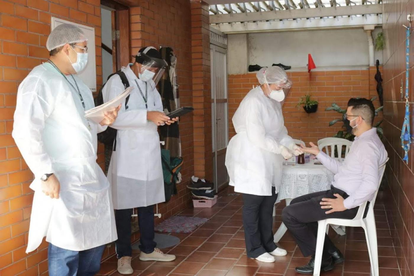 Homem está sentado fazendo teste rápido. Três pesquisadores estão em pé. #Paratodosverem