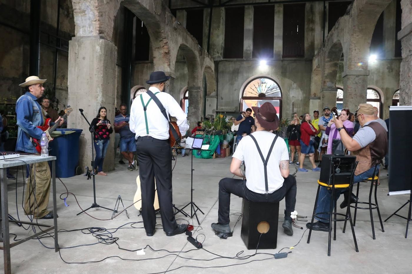 grupo musical está se apresentando em prédio antigo. Músicos estão de costas para a foto e público, de frente. #paratodosverem