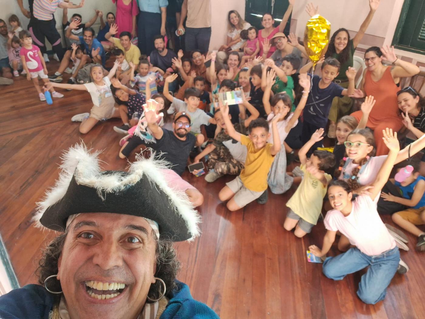 pirata em primeiro plano fazendo selfie com várias crianças ao fundo com os braços para cima. #paratodosverem 