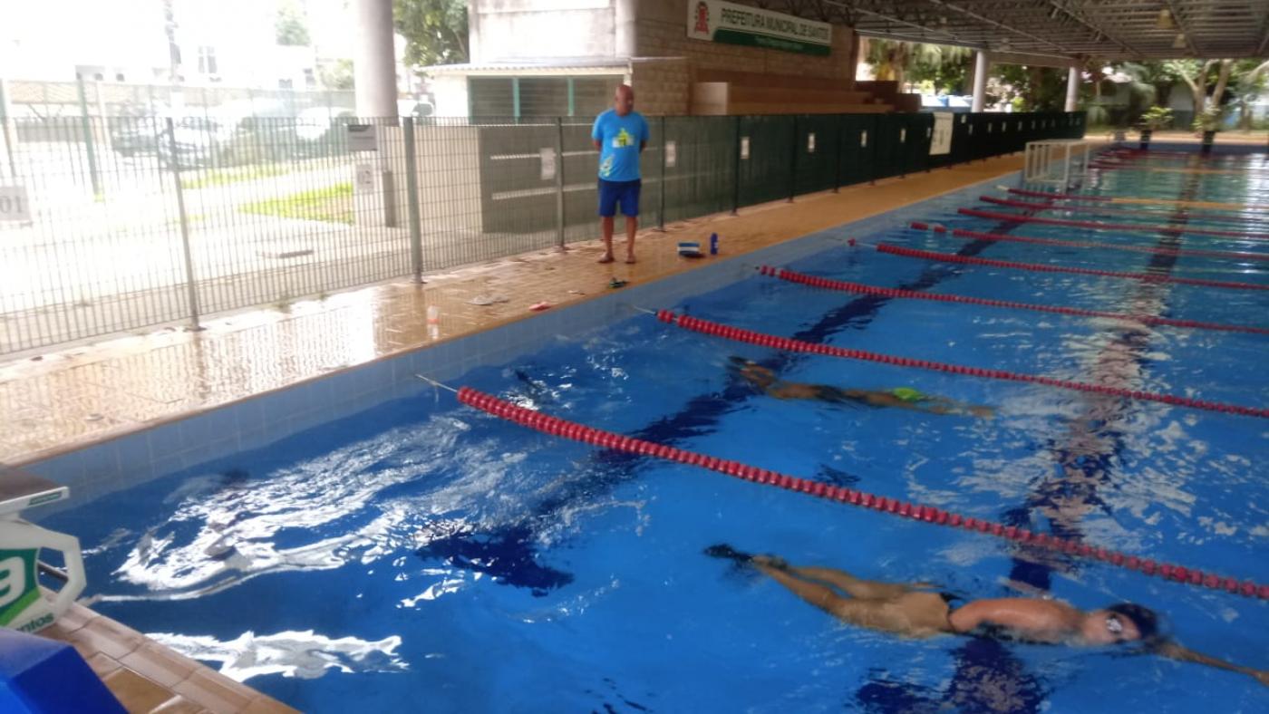 vista geral de piscina com pessoas nadando entre as raias. Homem, do lado de fora, está em pé. #paratodosverem