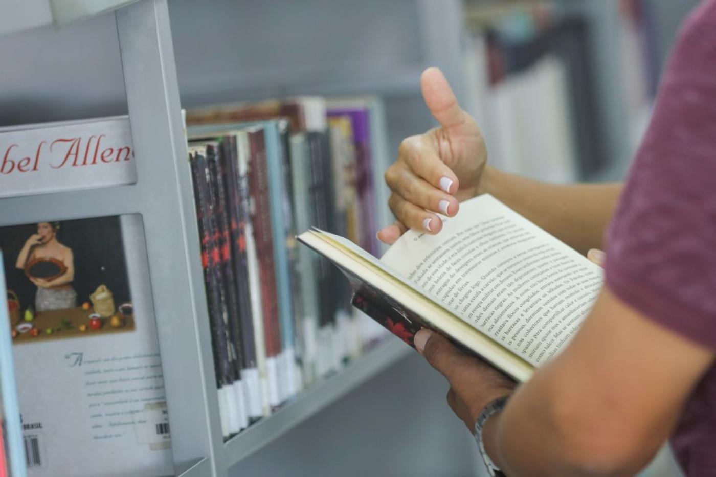 mãos seguram livro aberto. há livros numa estante ao fundo. #paratodosverem 