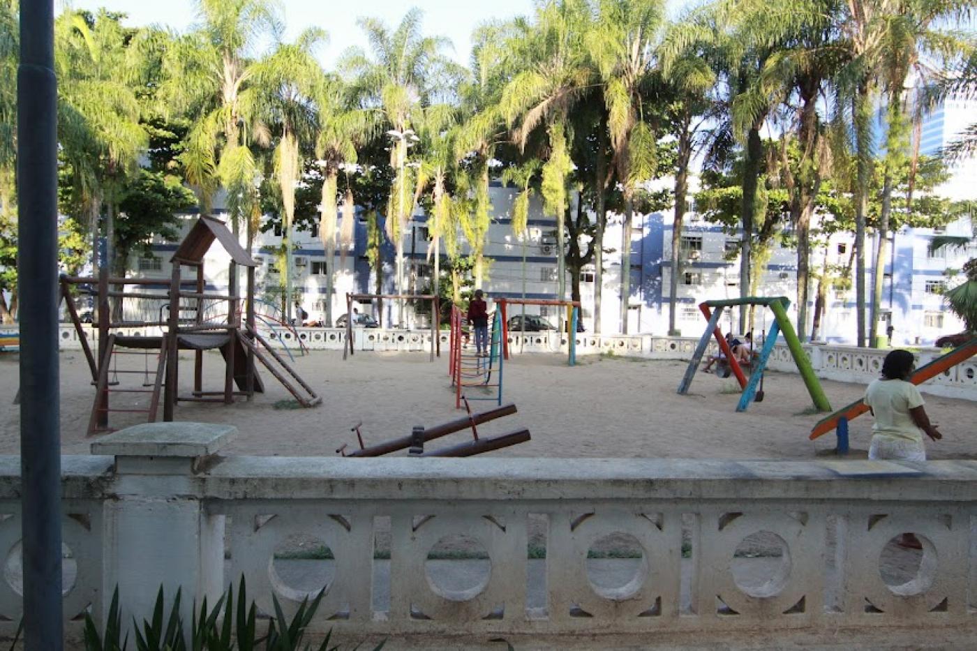 área de playground com brinquedos sobre piso de areia. À frente, o cercado é feito com as tradicionais muretas de santos. #paratodosverem