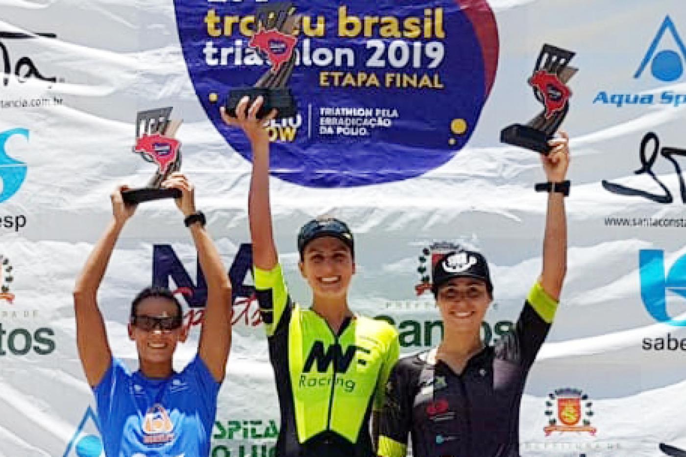 #pracegover Com Mariana Penatti ao centro, três mulheres com uniforme de competição erguem troféus no pódio