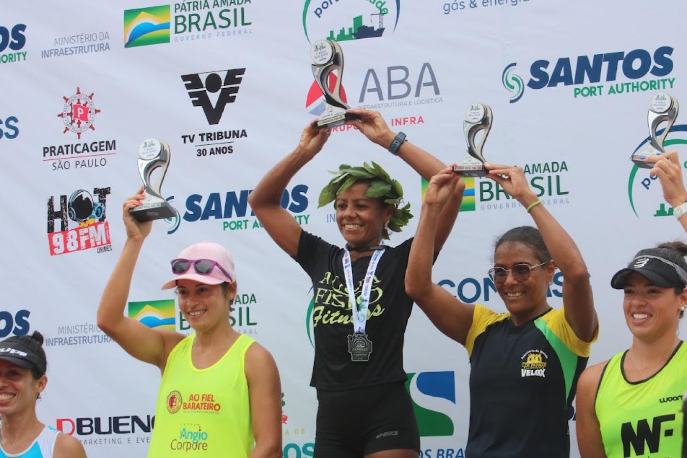 Pódio feminino com Ivanilda Pereira dos Santos ao centro. Elas estão todas com os braços para o alto segurando os troféus. #paratodosverem