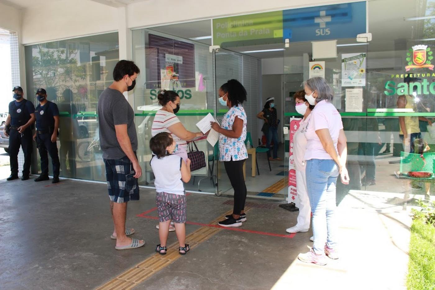 Casal e criança são recepcionados por profissionais na porta da unidade de saúde. #paratodosverem