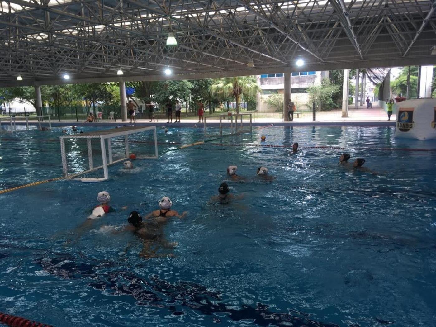 jovens disputam jogo na piscina. Do lado esquerdo e ao fundo se vê a trave sobre a água. #Paratodosverem