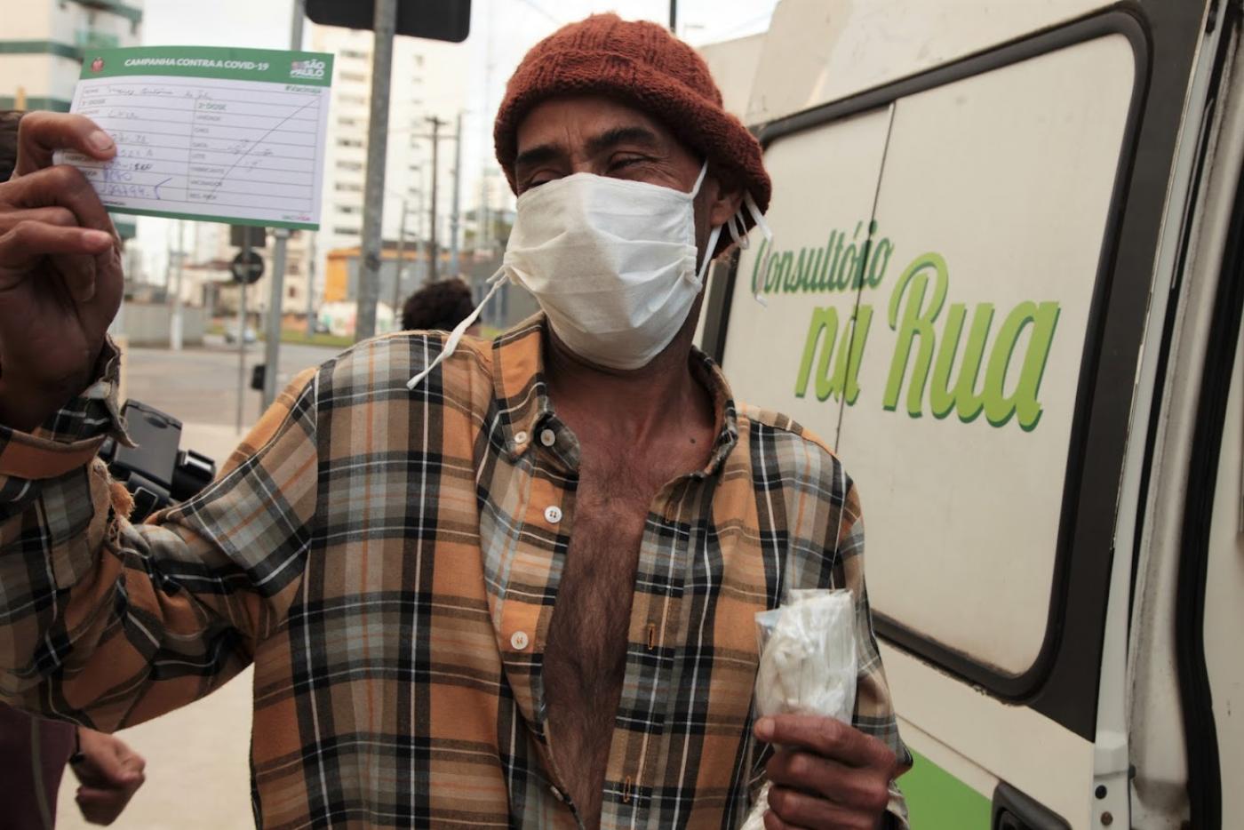 homem exibe cartão de vacinação. Ele está em pé ao lado de uma van onde se lê na porta; Consultório na Rua
