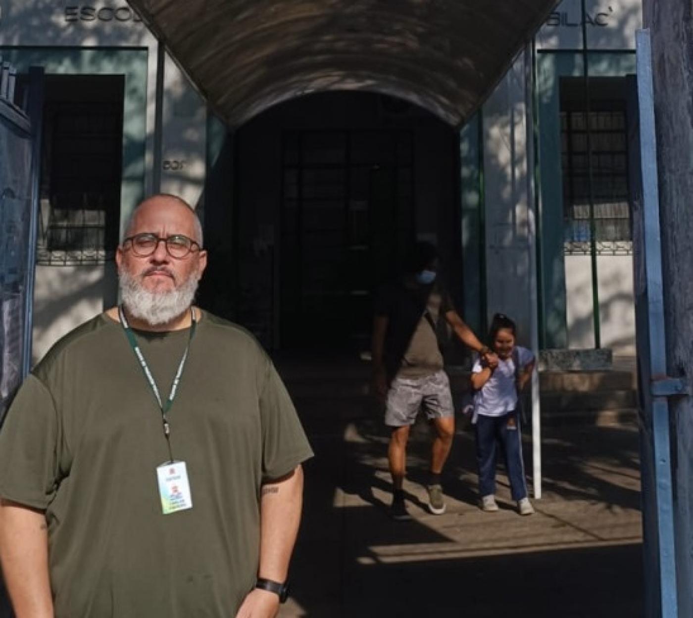 o porteiro Carlos na frente da escola. Há uma criança acompanhada do pai ao fundo saindo da unidade. #paratodosverem