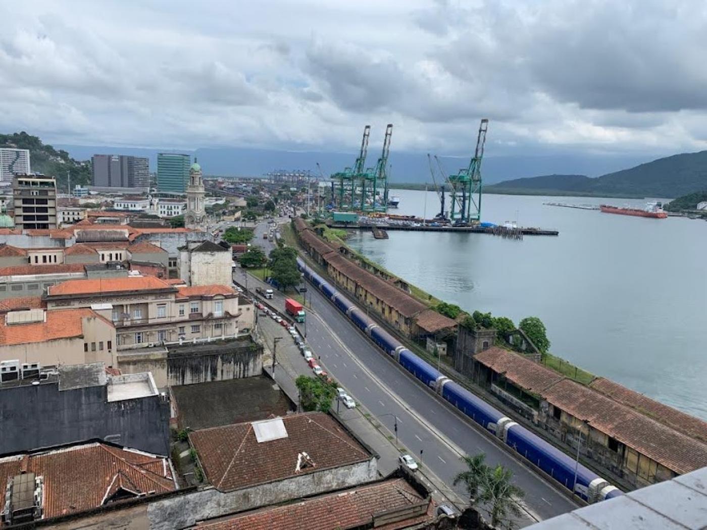 vista aérea da cidade ao lado esquerdo e o porto ao lado direito, como o mar à frente. Há portêineres ao fundo. #paratodosverem