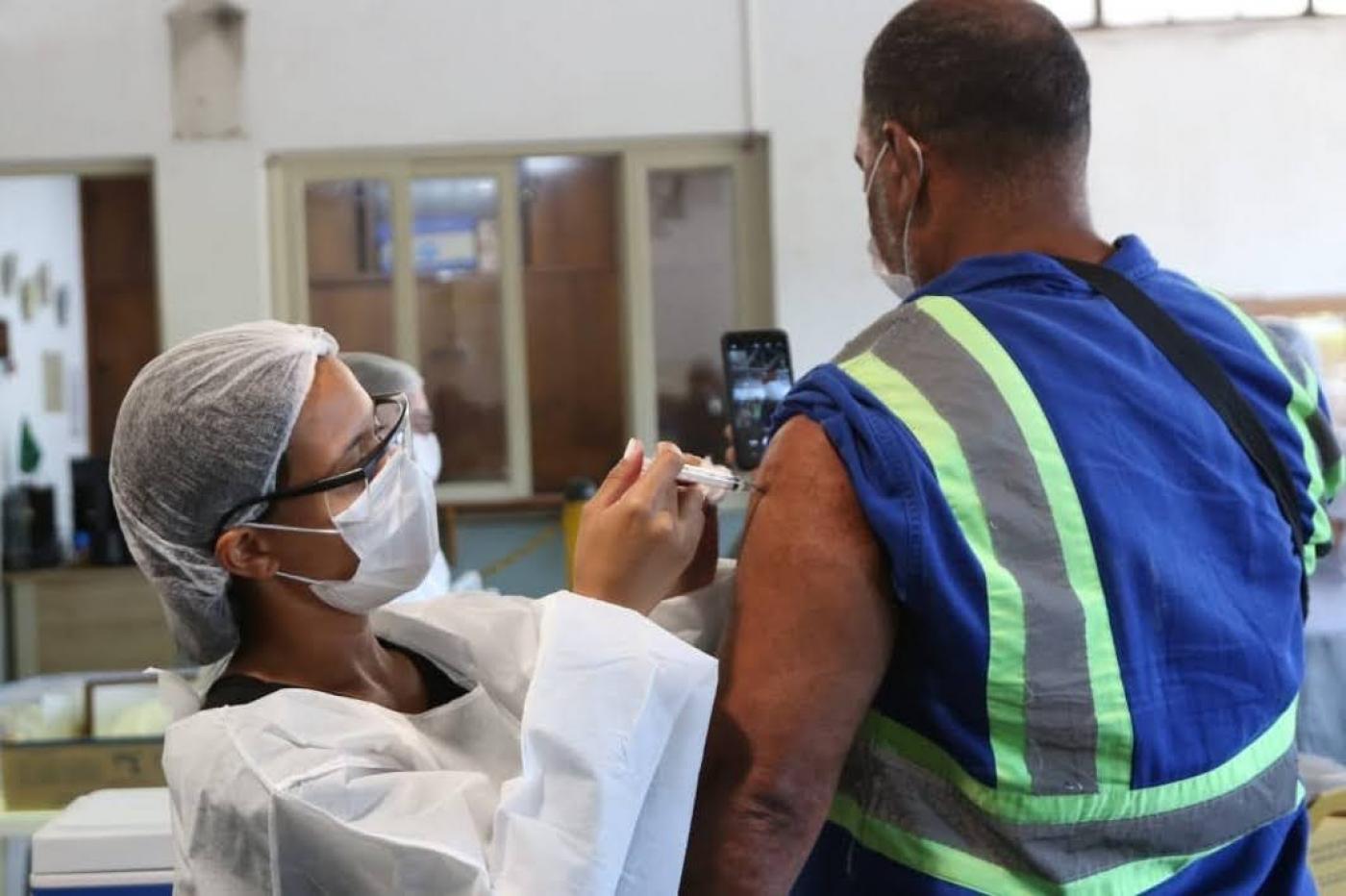 mulher vacina homem com roupa de operario e faixa reflexiva no uniforme. #paratodosverem