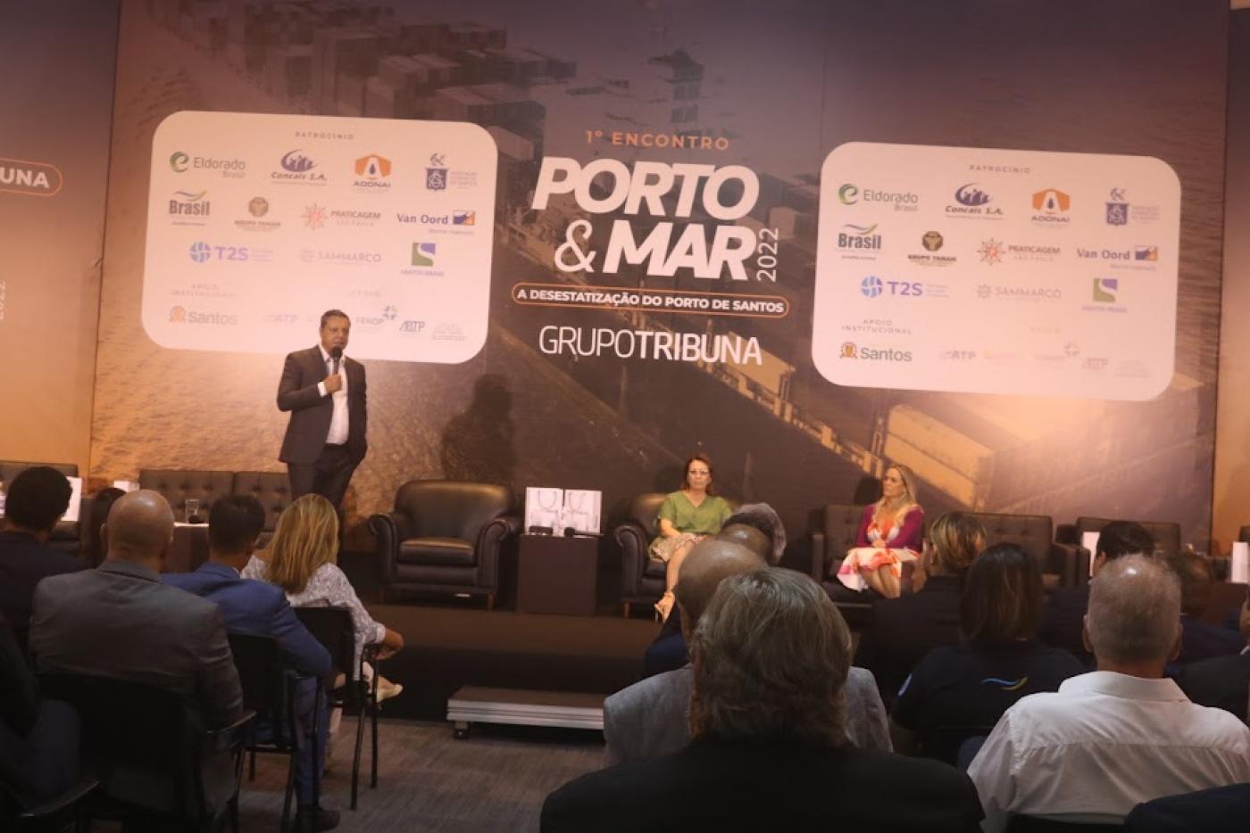 prefeito fala em palco. Ele está ao lado esquerdo da foto em pé. Do lado esquerdo, duas mulheres sentadas. A plateia assiste. Ao fundo, um painel onde se lê Porto & Mar. #paratodosverem