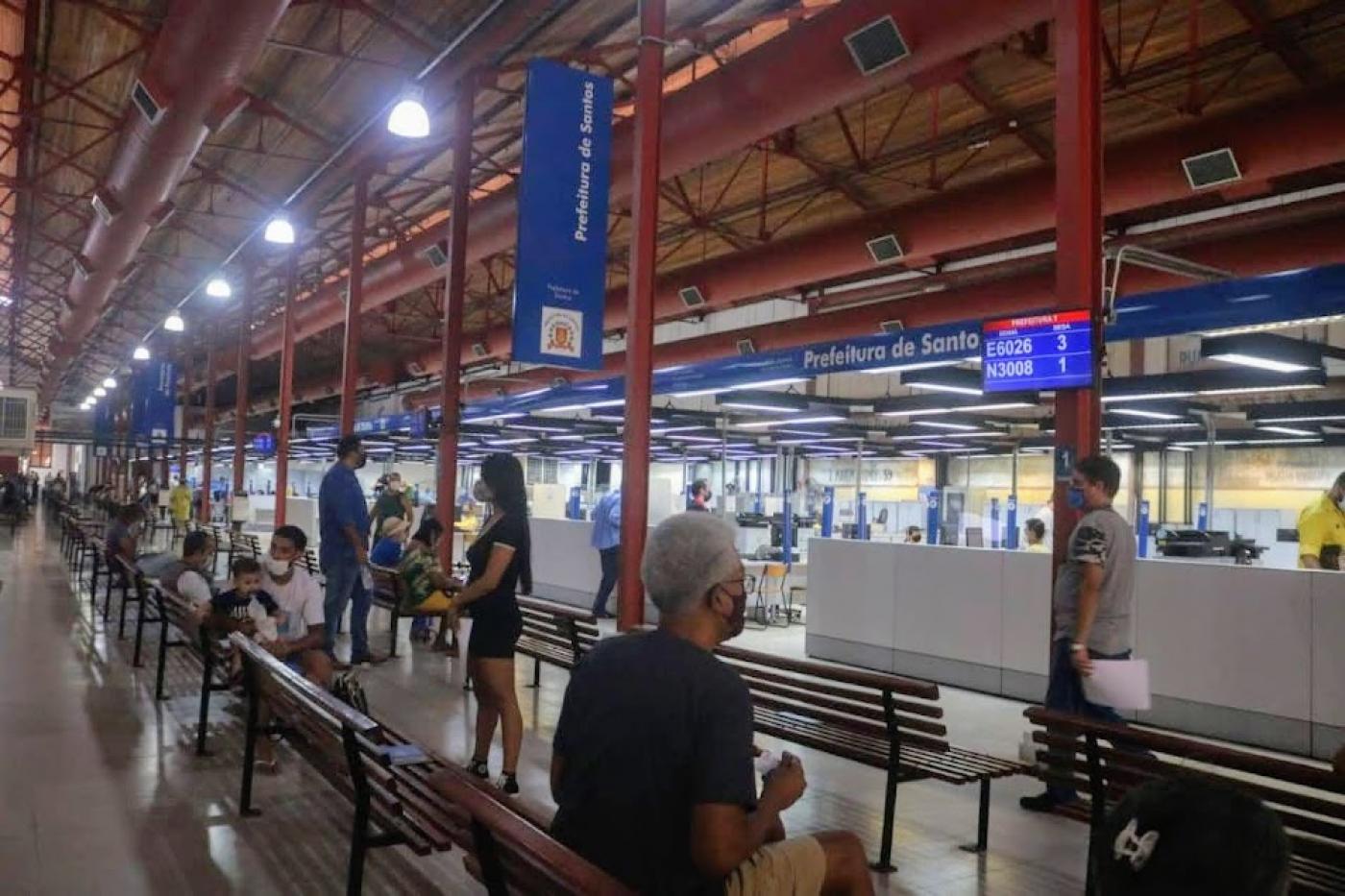 unidade do poupatempo, com pessoas sentadas em bancos. Ambiente é um antigo prédio restaurado, com estruturas aparentes e paineis indicando on